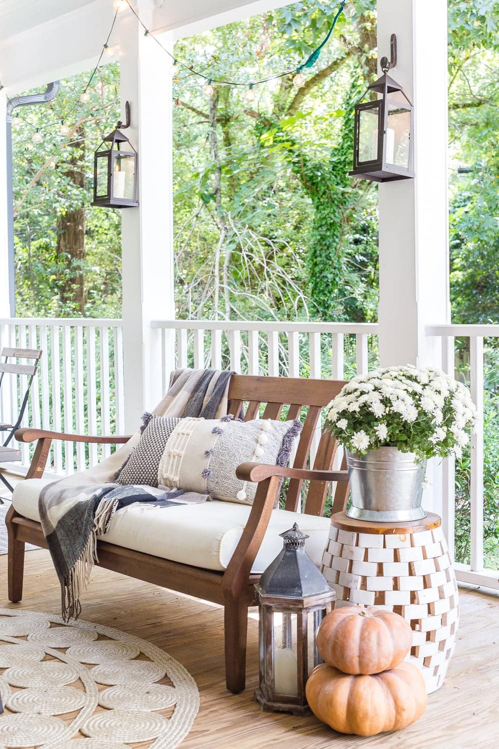 cozy fall porch with plaid throw blankets, textured pillows, pumpkins, lanterns, mums, and string lights