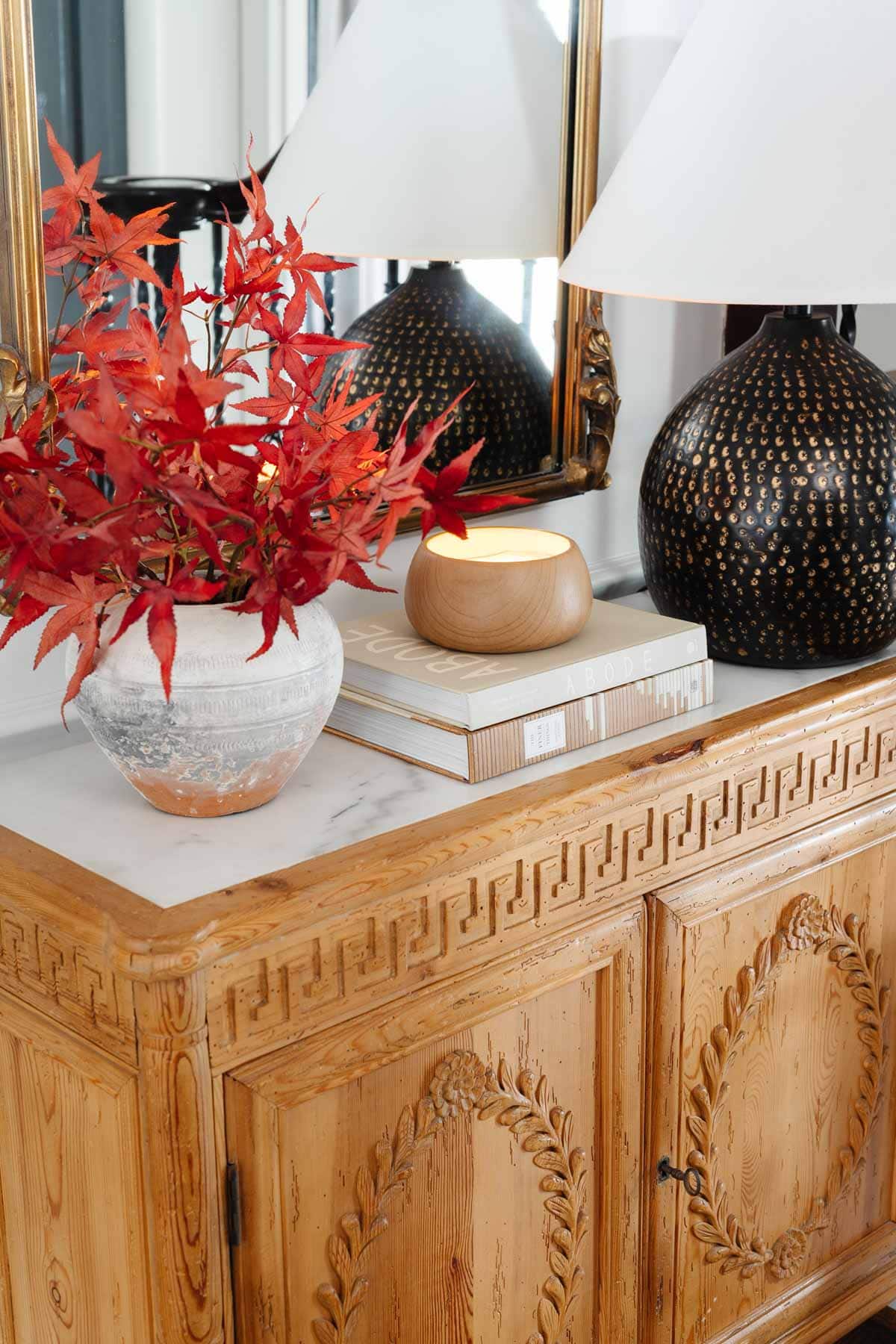 carved wood cabinet in foyer with vase of fall leaves, gold ornate mirror, and black lamp
