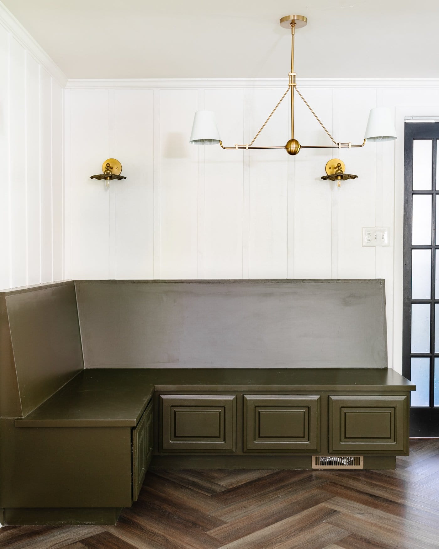 breakfast nook with green banquette bench, white batten wall made of lattice strips, and brass lighting